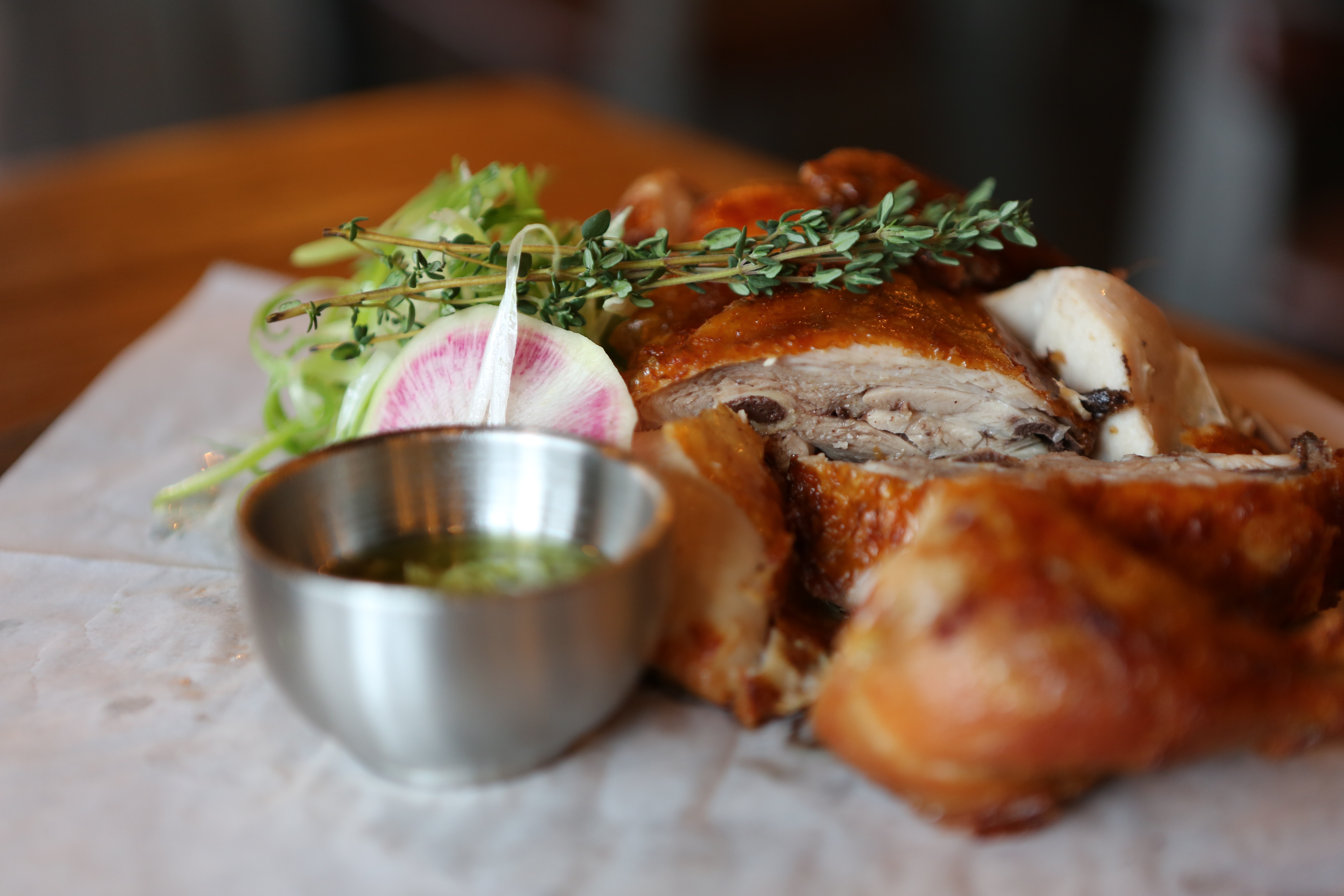 Roasted chicken on a table with herbs and a small silver bowl.