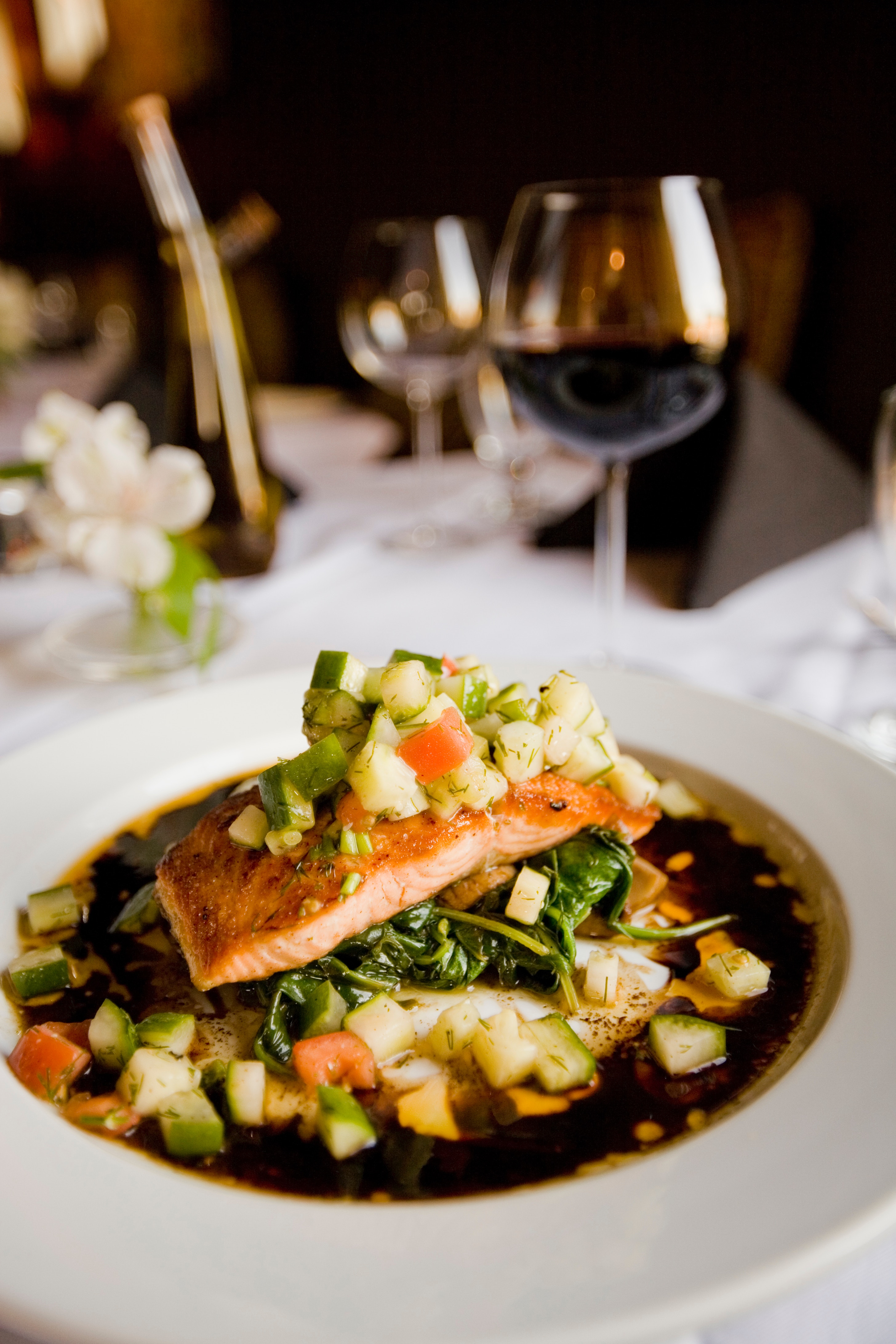 Plate of cooked fish with garnishes on white tablecloth and glass of red wine in background