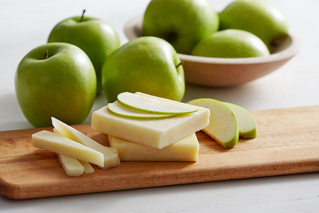 White background with wooden cutting board, granny smith apple slices and white cheese slices. Green granny smith apples in background.