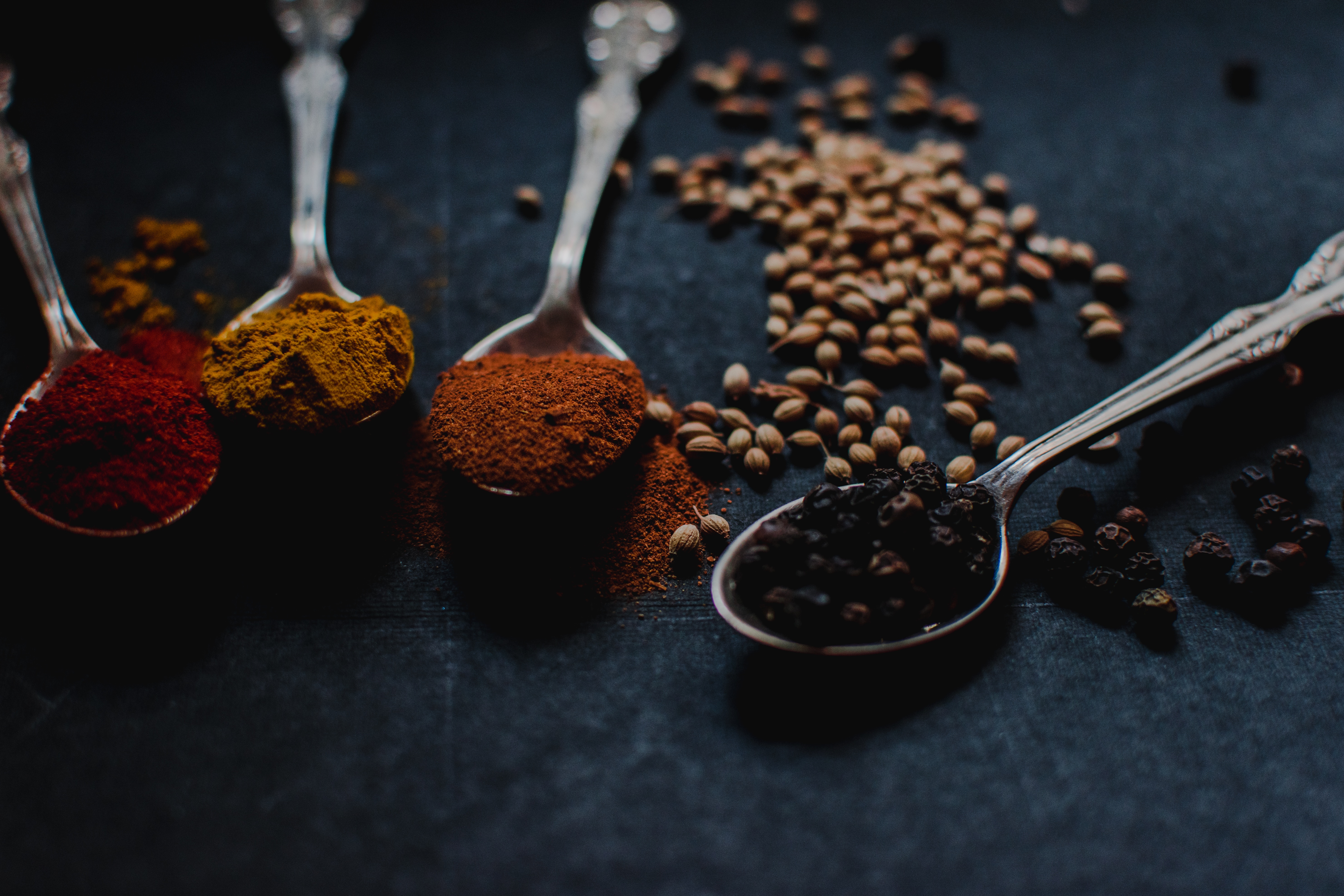 Four metal spoons filled with spices against black surface, spilled spices on surface.