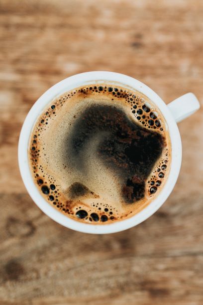 Looking down at a frothy cup of black coffee in a white mug (against wooden table)